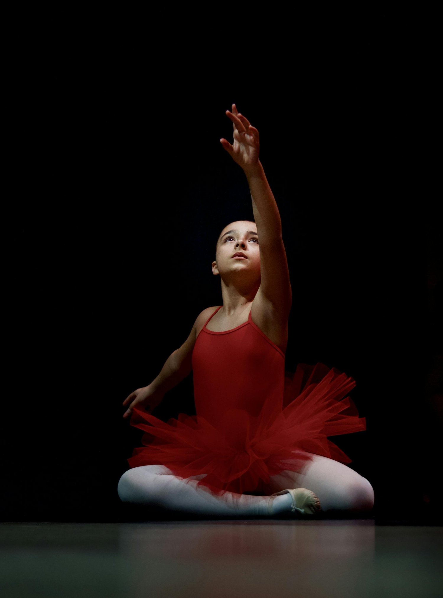 A young girl in a red ballet costume sitting in a ballet pose with one arm raised.