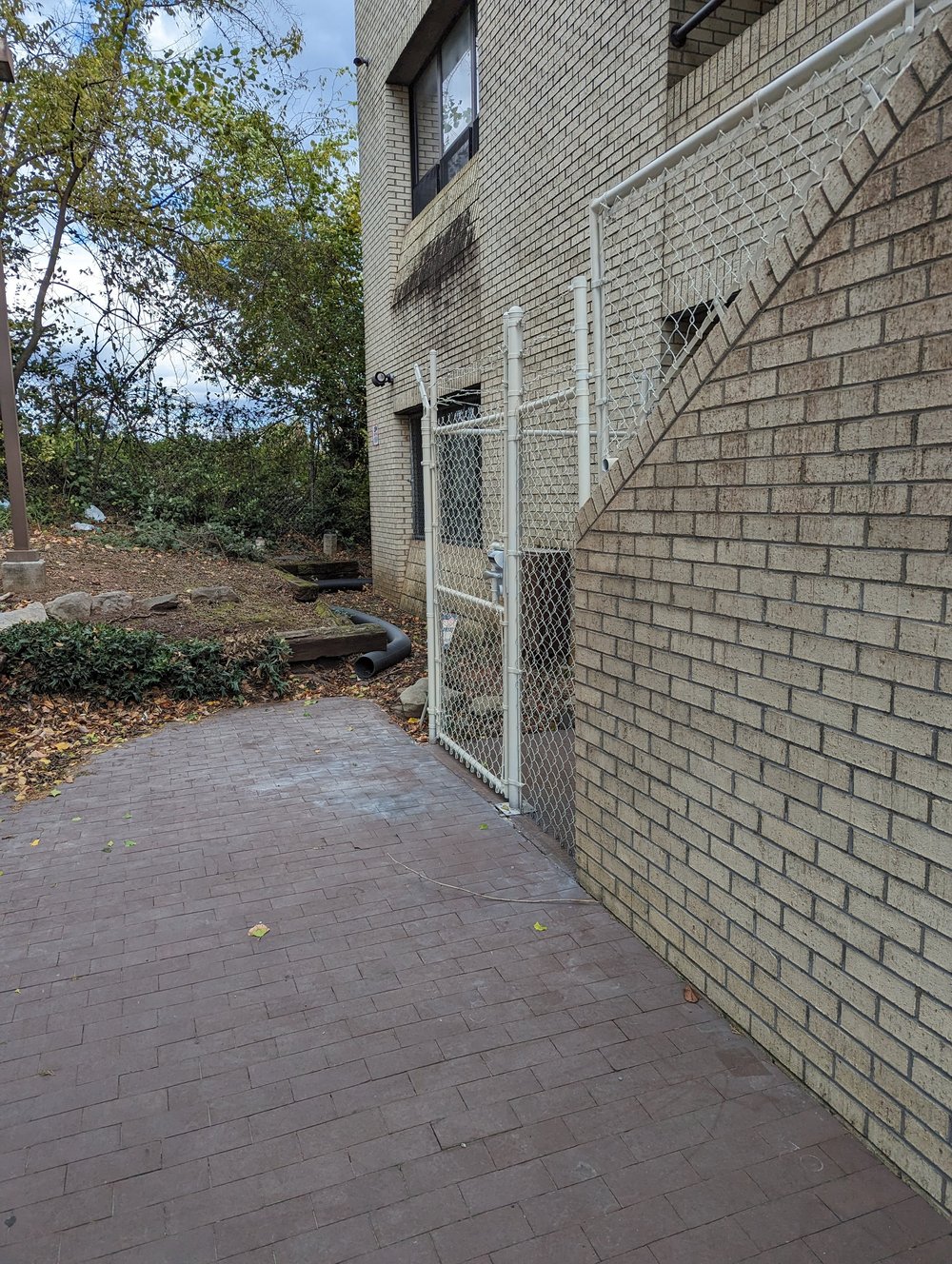 A brick walkway along the left side of the building leading to a white chain-link gate under an outdoor stairway.