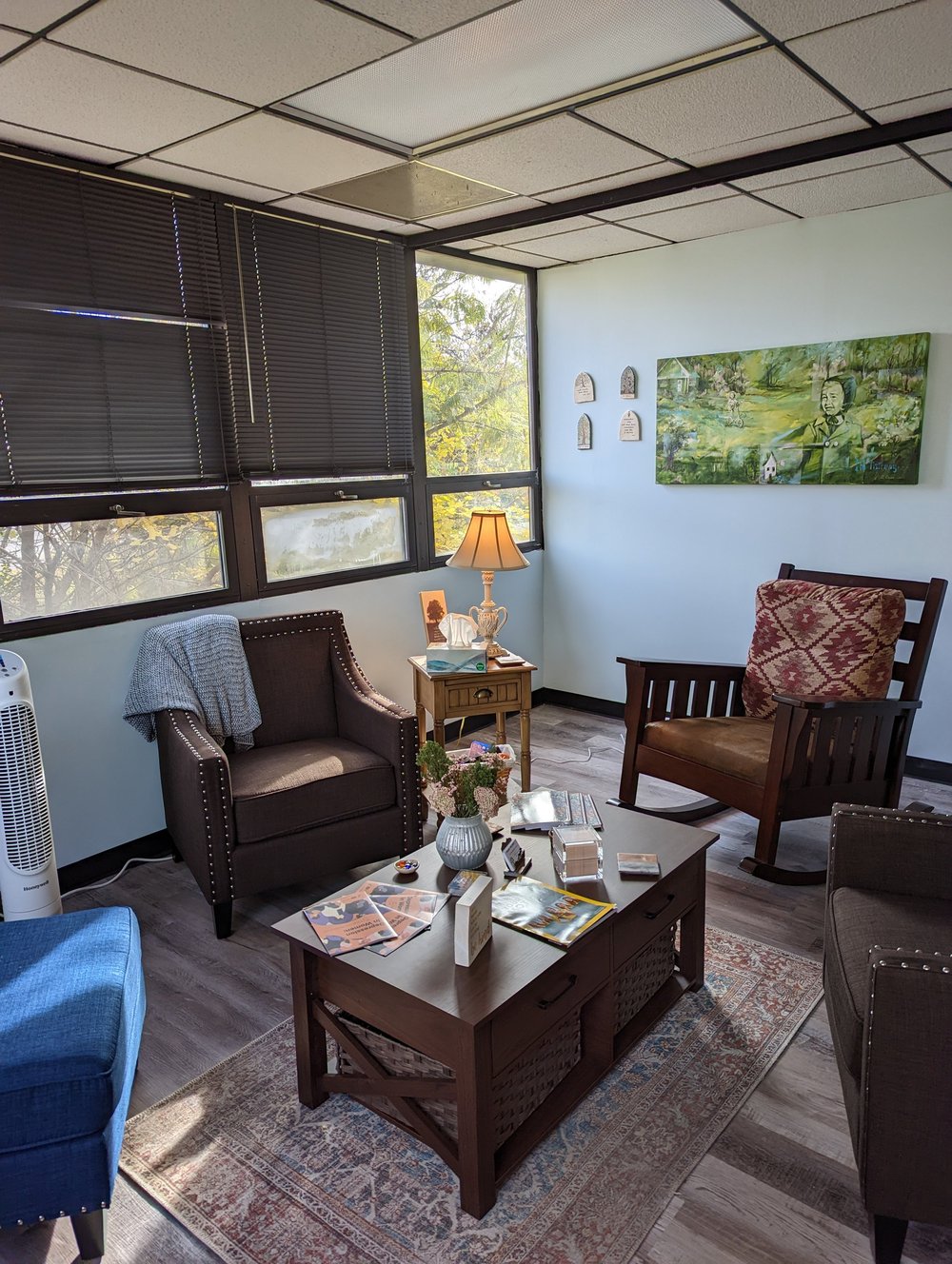 A counseling room at Trauma Wellness with armchairs, a rocking chair, a lamp and a large green painting.